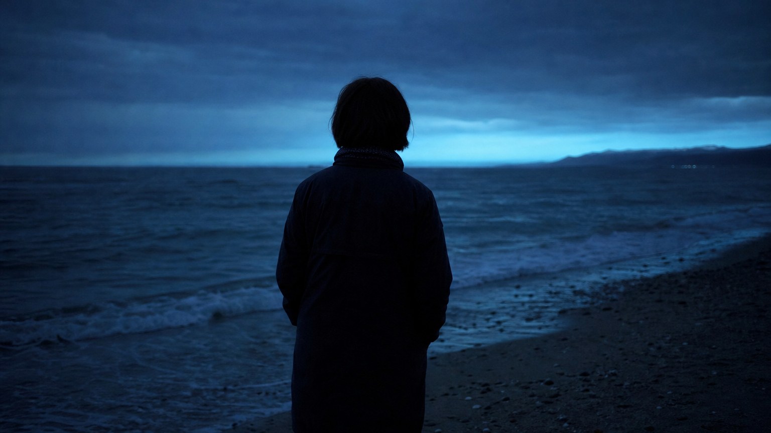 夕暮れの浜辺で海を見つめる人物の後ろ姿（明日への予告ビジュアル）